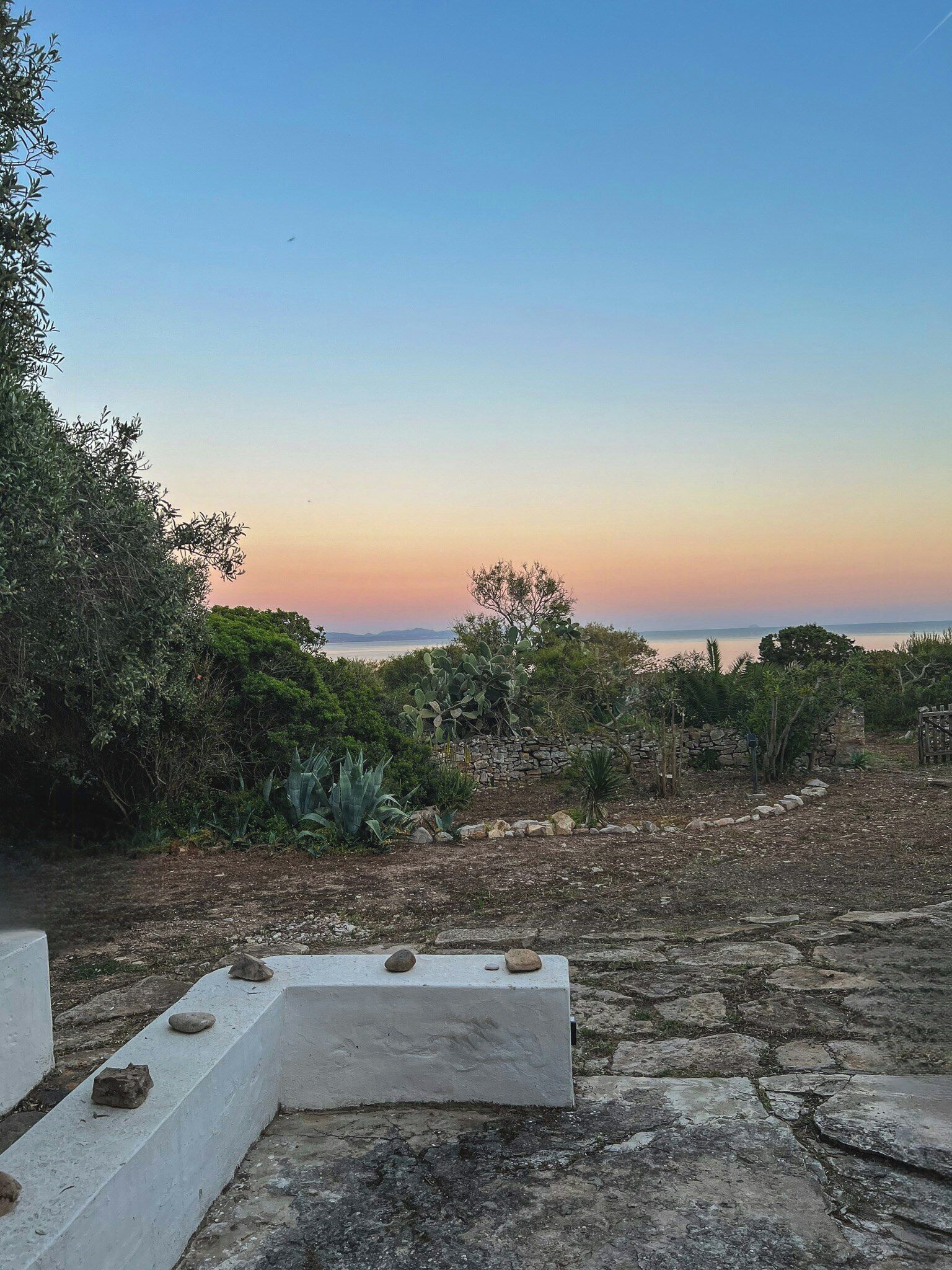 A stone pathway leading to a seating area with a view of the sea at sunset.