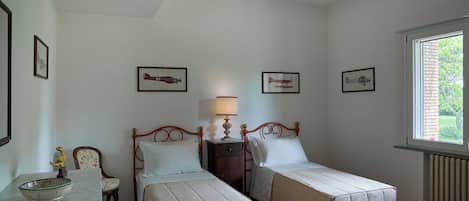 A bedroom with two single beds, a bedside table with a lamp, a small table with a decorative bowl, and framed pictures on the wall.