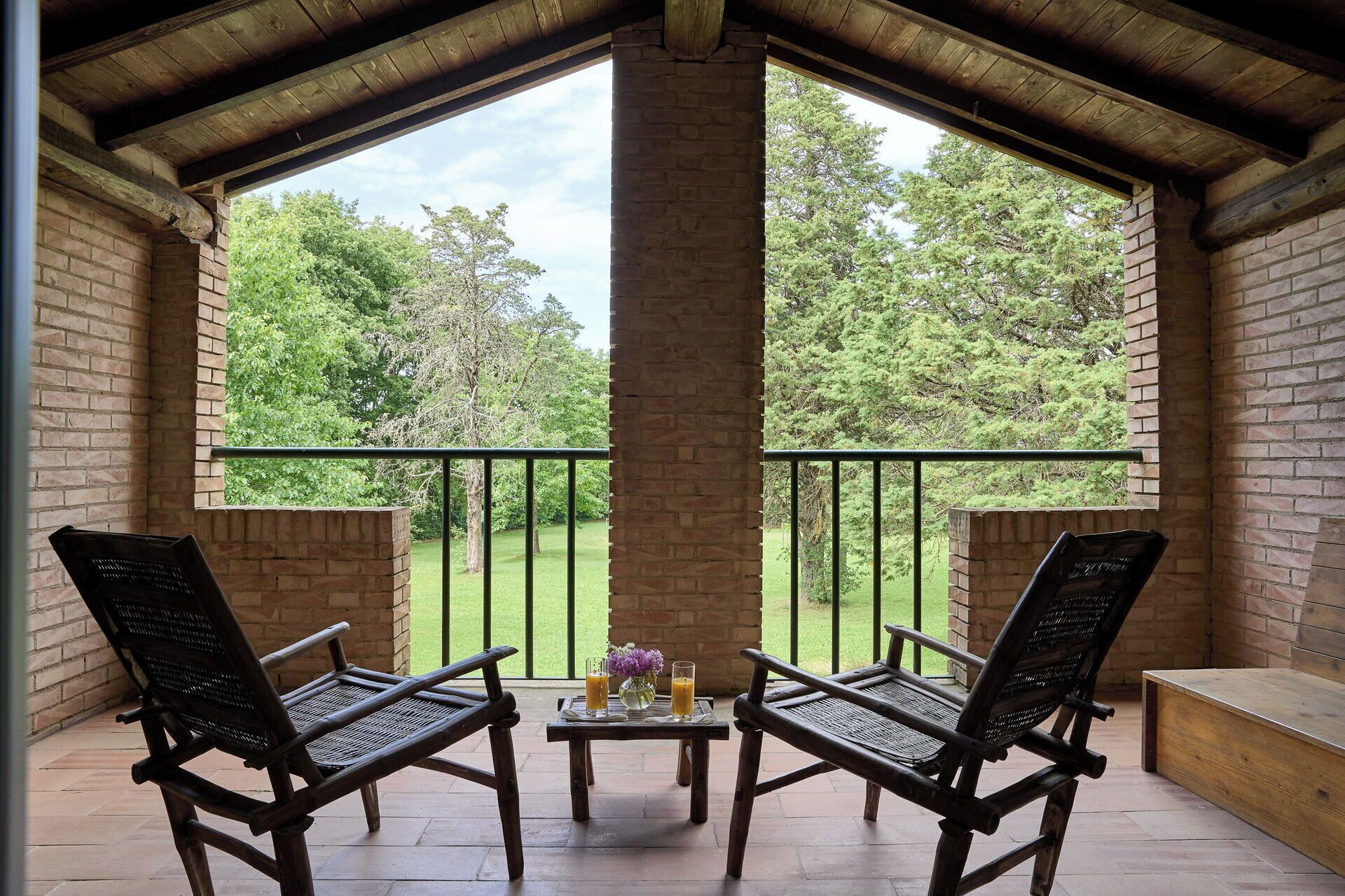 Een overdekte buitenruimte met twee stoelen, een tafel, een bakstenen pilaar en uitzicht op een groen landschap.