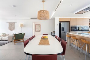 Eine moderne Küche mit Edelstahlgeräten, ein Essbereich mit weißem Tisch und ein Wohnbereich mit grünem Sessel und Wandbehang aus Makramee.
