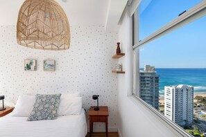 Ein Schlafzimmer mit einem Bett, einem Nachttisch, einer Lampe, einer Wandablage und Blick auf das Meer und Gebäude.