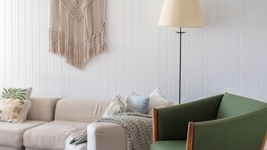 Een woonkamer met een bank, een groene fauteuil, een vloerlamp en een macramé wanddecoratie.