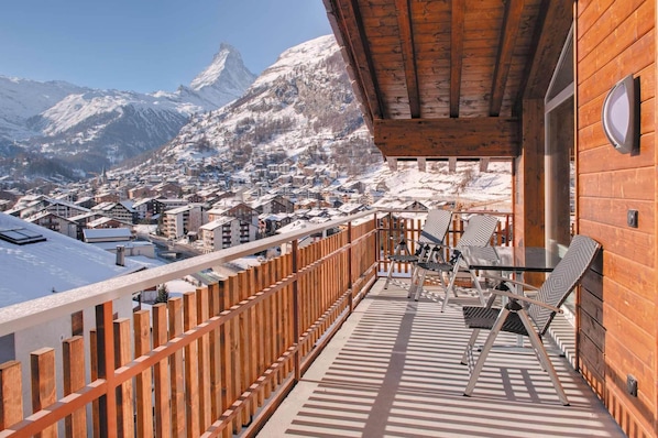 A wooden balcony with a view of a snow-covered mountain and a town below. - Powder Snow Loft - by Zermatt Apartment Rentals : Matterhorn Views, sleeps 6-7 (zermatt)