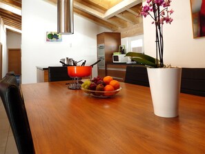 A dining area with a wooden table, a bowl of fruit, a white vase with a plant, and a kitchen in the background. - Powder Snow Loft - by Zermatt Apartment Rentals : Matterhorn Views, sleeps 6-7 (zermatt)