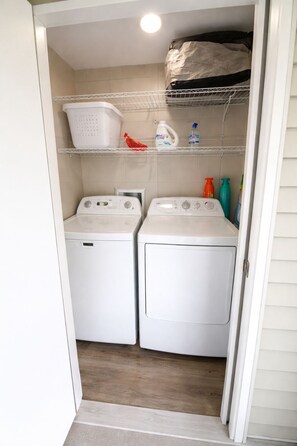A laundry room with two white front-loading washing machines, a shelf with laundry detergent, and a basket. - LUXURY 1-bedroom condo in Destin (Destin)