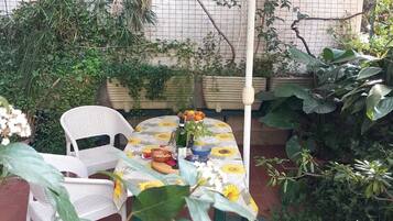 Ein kleiner Außenbereich mit Sitzgelegenheit, einem Tisch mit einem sonnenblumenmuster-Tischdecke, umgeben von üppigem Grün und einem weißen Stuhl.