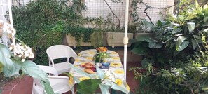 Un pequeño espacio al aire libre con una mesa cubierta por un mantel estampado con motivos de girasoles, rodeado de abundante vegetación y una silla blanca.