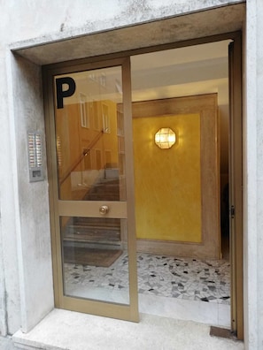 A building entrance with a glass door marked with a 'P' for parking, a staircase inside, and a wall-mounted light fixture. - Casa Dino room (Roma)
