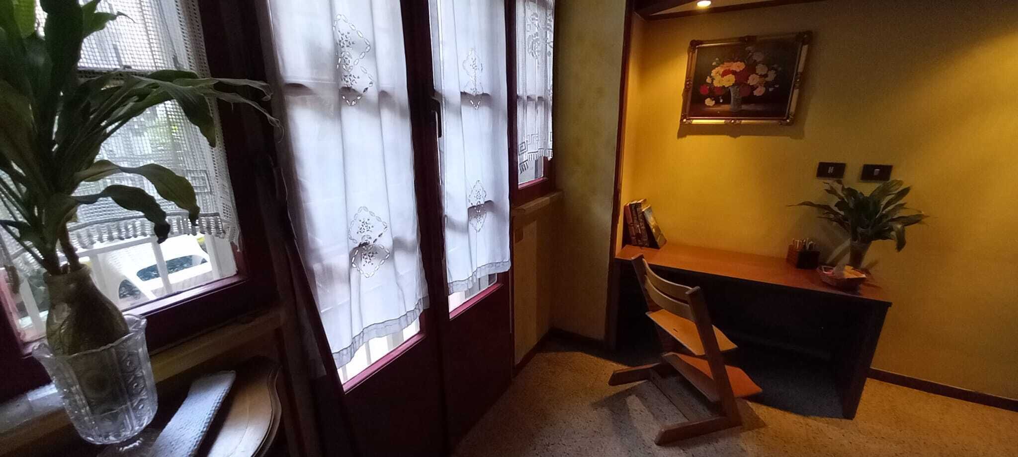 Ein Eckzimmer mit Schreibtisch aus Holz, einem Stuhl, einer Zimmerpflanze und einem Fenster mit Spitzenvorhängen.