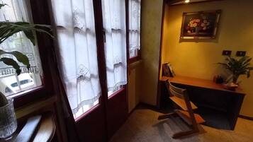 Ein Eckzimmer mit Schreibtisch aus Holz, einem Stuhl, einer Zimmerpflanze und einem Fenster mit Spitzenvorhängen.