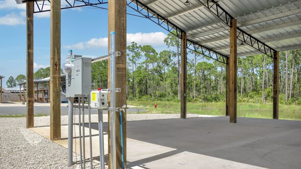 A covered outdoor area with a concrete floor, metal poles, and a utility box labeled '156 SIMMONS WAY'. - RV Site, Full Hookup, Flo's RV Home (Port St. Joe)
