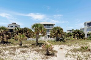 Une propriété située en bord de mer, avec une maison principale dotée d’un balcon, entourée de palmiers et bordée d’un paysage sablonneux.