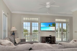 Un salon doté d’un ventilateur de plafond, d’un téléviseur diffusant une scène de plage et d’une vue sur un balcon extérieur, accessible par une porte vitrée.