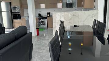 Et moderne køkken med stålemmer, et spiseområde med et glasbord og sorte stole, samt en opholdsstue med en sort sofa.
