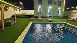 A pool area with a house in the background, a pergola, and a well-lit exterior.