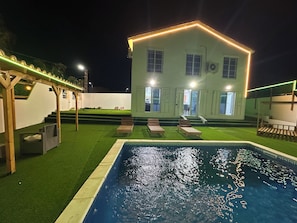 A pool area with a house in the background, a pergola, and a well-lit exterior.