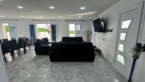 A spacious living area with a large black sofa, a flat-screen TV mounted on the wall, and a ceiling with recessed lighting.