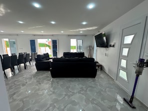 A spacious living area with a large black sofa, a flat-screen TV mounted on the wall, and a ceiling with recessed lighting.