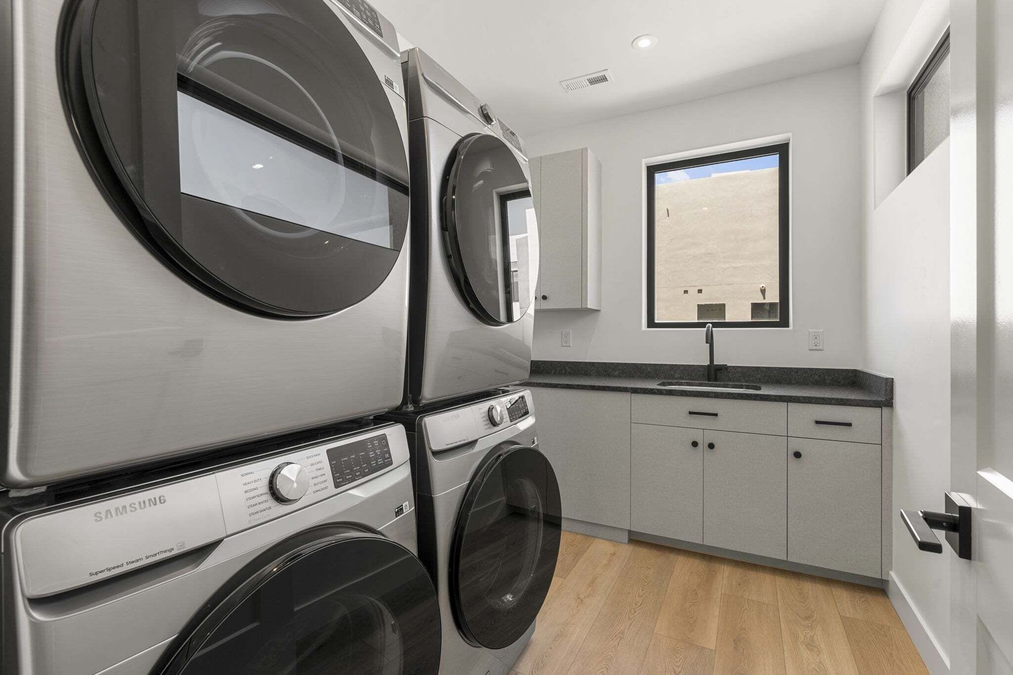 A compact laundry room with stacked washers and dryers, a small sink, and a window with a view.