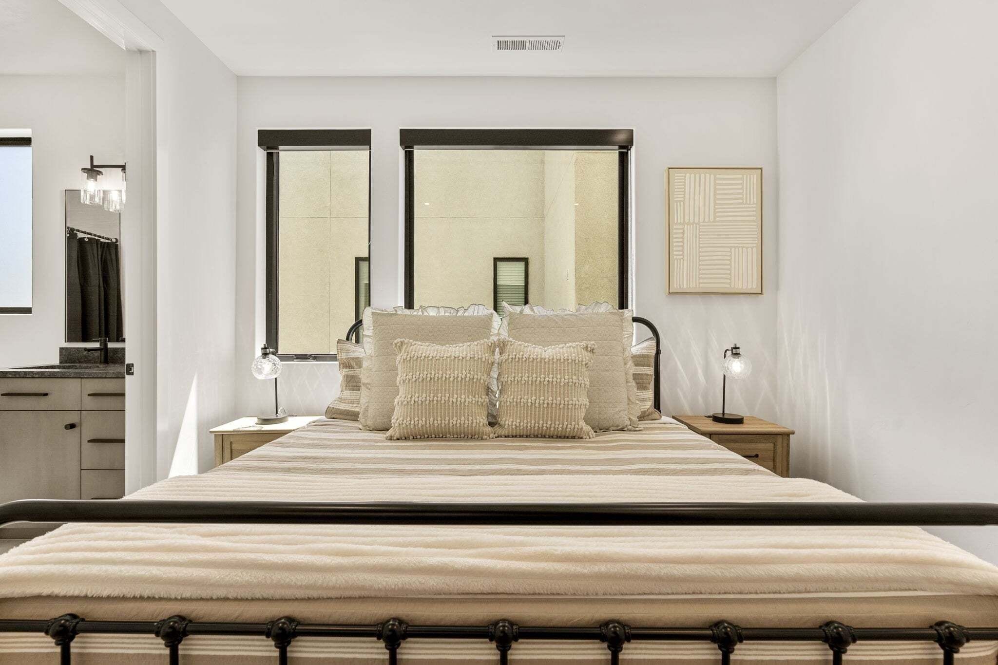 A bedroom with a large bed, two bedside tables, a mirror, a window, and a wall-mounted artwork.