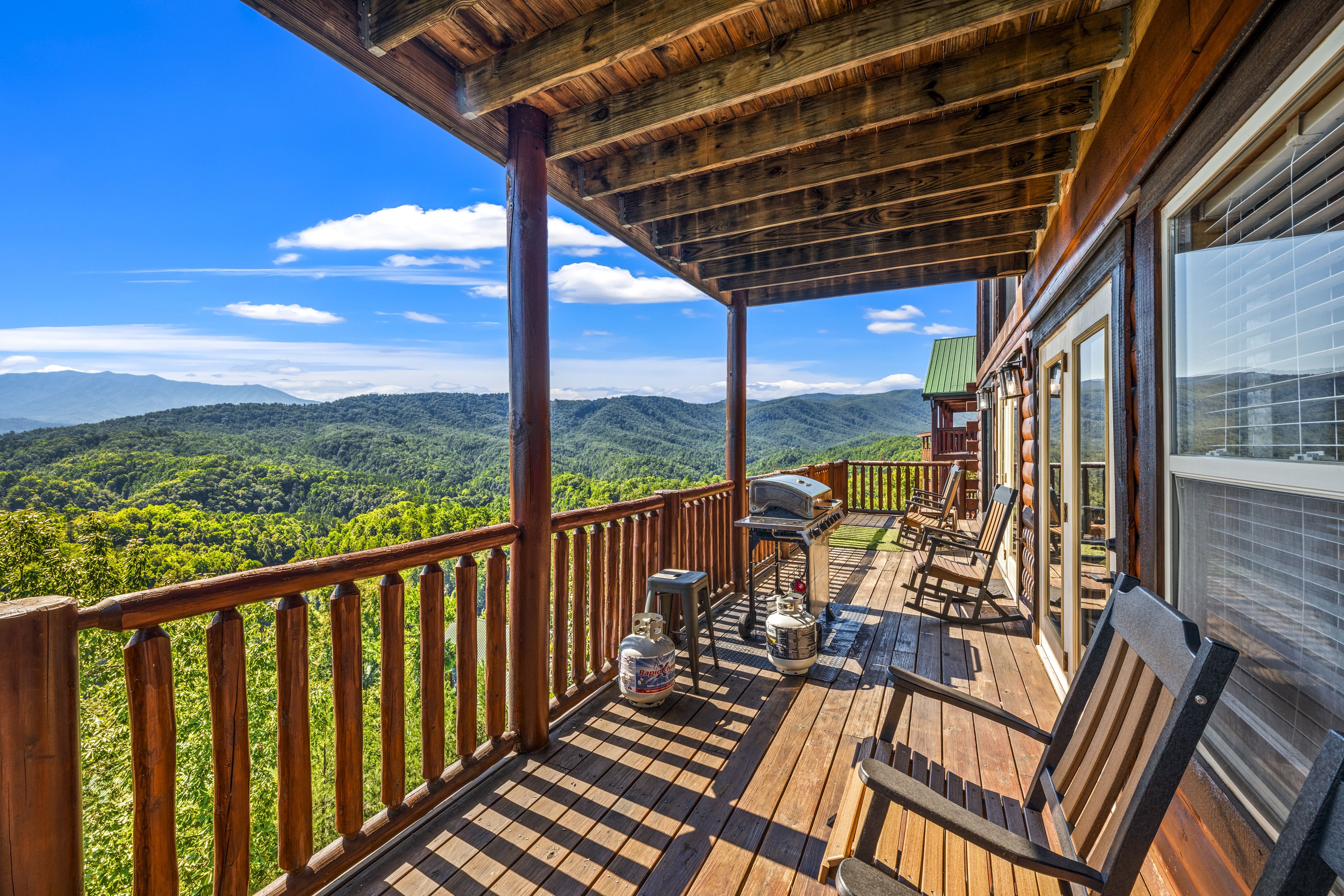 Una terraza de madera con vistas a una cadena montañosa boscosa, una parrilla y asientos exteriores.