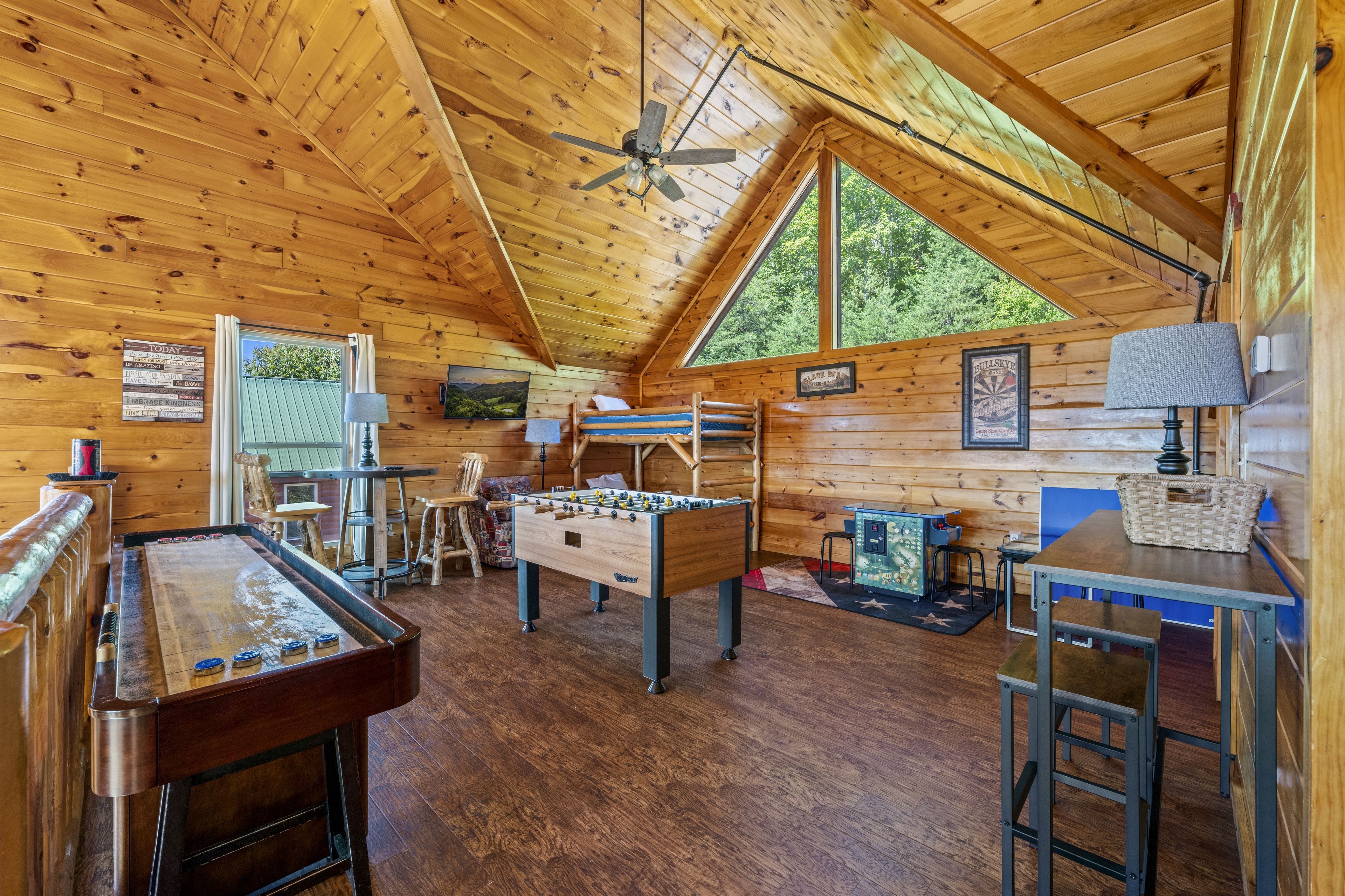 A cozy room with wooden walls, a ceiling fan, a foosball table, a pool table, and a window with a view of trees.