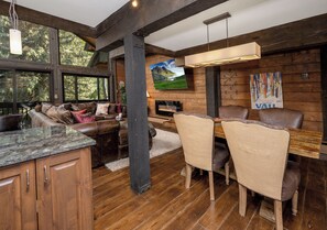 Ein rustikales Interieur mit einem Holztisch, Ledersesseln, einem Kamin und einem großen Fenster mit Blick auf Grünflächen.