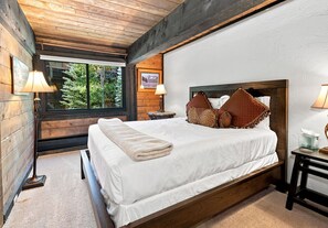 Ein Schlafzimmer mit einem Holzkopfteil, einem großen Bett mit weißer Bettwäsche, zwei Nachttischlampen und einem Fenster mit Blick auf Bäume.