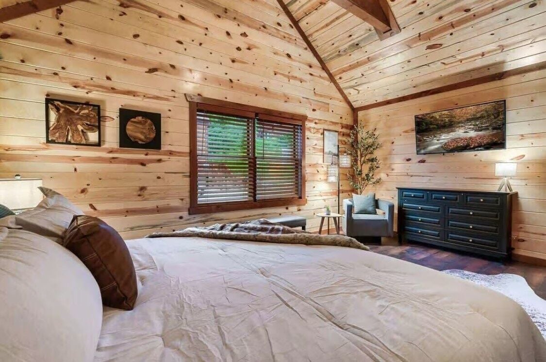 A wooden cabin bedroom with a bed, a dresser, a chair, and a window with blinds.