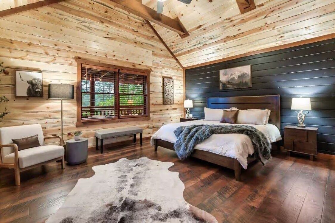 A log cabin bedroom with a bed, a bench, a cowhide rug, and a window with blinds.