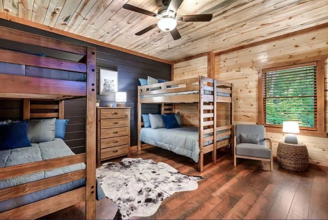 A cabin interior with bunk beds, a ceiling fan, a wooden floor, and a window with blinds.