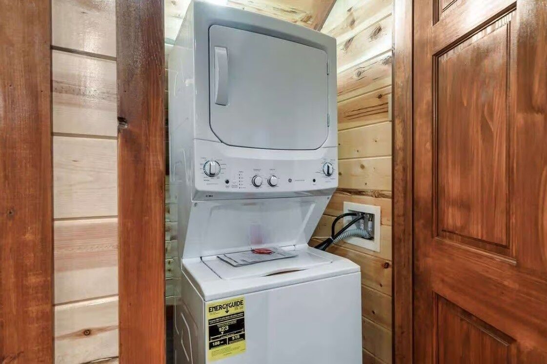 Lavadora blanca compacta con perillas de control y pantalla digital, ubicada en una cabaña de madera con una puerta y un enchufe a la vista.