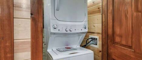 Lavadora blanca compacta con perillas de control y pantalla digital, ubicada en una cabaña de madera con una puerta y un enchufe a la vista.