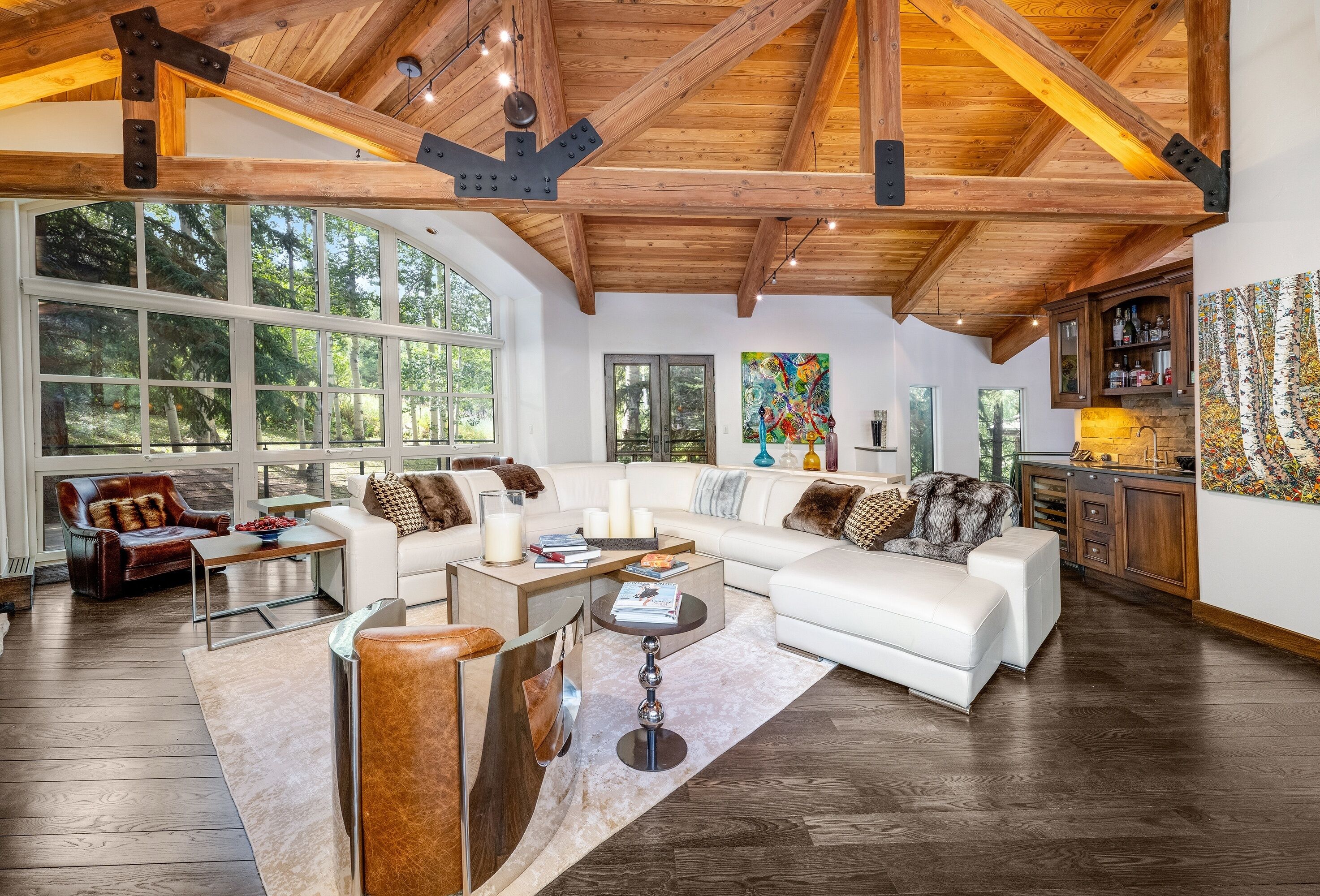 Un salon spacieux avec un grand canapé d’angle, une table basse et une cheminée. De grandes fenêtres laissent entrer la lumière naturelle et offrent une vue sur la forêt environnante.