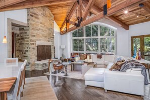 A spacious living room with a stone fireplace, a large window with a view of trees, and a white leather sofa. - For486 5 Bedroom Home (Vail)