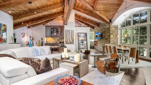 A spacious living area with a high ceiling, wooden beams, a fireplace, and a large window overlooking greenery. - For486 5 Bedroom Home (Vail)