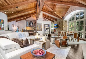 A spacious living area with a high ceiling, wooden beams, a fireplace, and a large window overlooking greenery. - For486 5 Bedroom Home (Vail)
