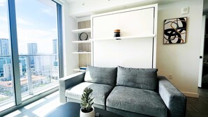 A modern living room with a grey sofa, a potted plant, and a city view through large windows. - 17th floor skyline retreat in Downtown Miami rooftop pool, coworking lounge and self check-in (Miami)
