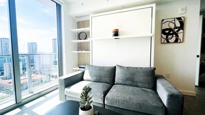 Ein modernes Wohnzimmer mit einem grauen Sofa, einer Topfpflanze und Blick auf die Stadt durch große Fenster.