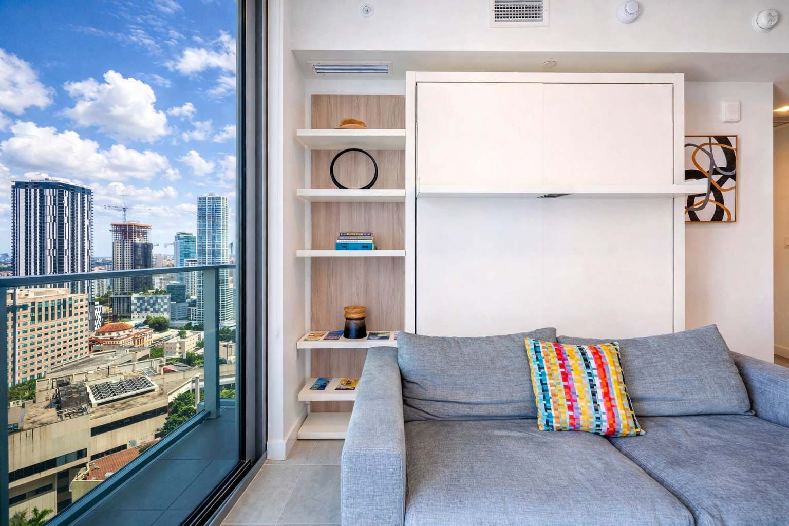 A modern living room with a grey sofa, colorful cushions, and built-in shelves. A large window offers a city view.