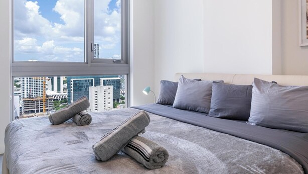 A bedroom with a large bed, a view of the city through a window, and a cityscape visible outside. - 17th floor skyline retreat in Downtown Miami rooftop pool, coworking lounge and self check-in (Miami)