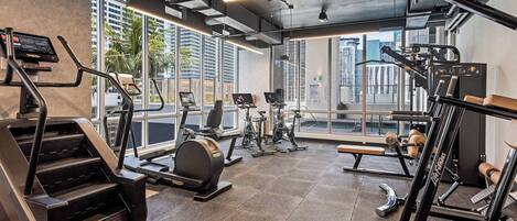 A modern gym with various exercise equipment, large windows, and a city view.