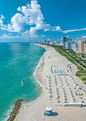 A beach with rows of umbrellas and lounge chairs, a lifeguard tower, and a city skyline in the background. - Skyline Studio w/ Pool & Gym | Walk to Bayside (Miami)