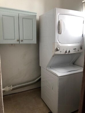 A compact laundry room with a front-loading washing machine and a dryer, a light blue cabinet above, and a visible hose. - P. H. PRIVATE PATIO W/ THERMO TUB./POOL, 1 BLK TO RESTRNT & MRKT, CLOSE TO BEACH (Playa del Carmen)