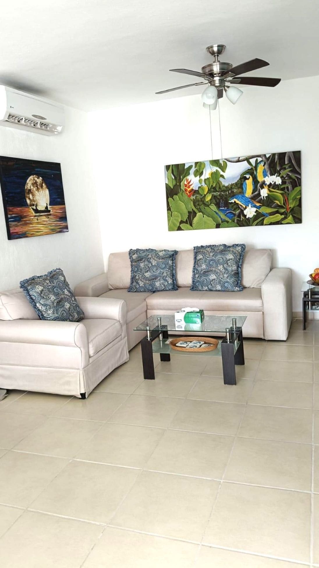 A living room with a sofa, armchairs, a coffee table, a ceiling fan, and two wall-mounted paintings.