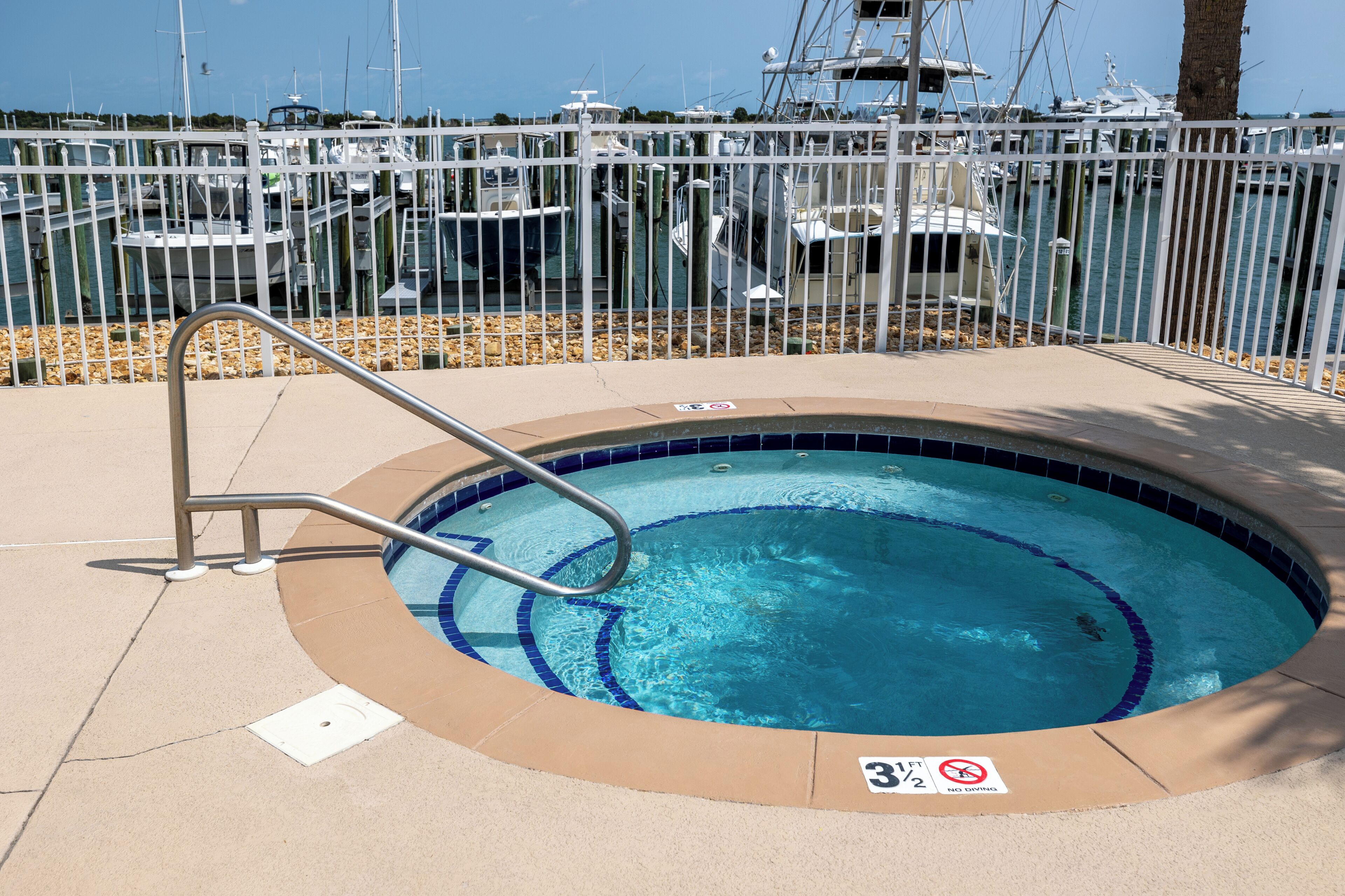 Une petite piscine avec un escalier en métal, entourée d’une clôture blanche, et des bateaux amarrés en arrière-plan.