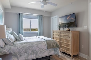 Ein Schlafzimmer mit Bett, Kommode, Deckenventilator, Fenster mit Vorhängen und einem flachbildfernseher an der Wand.