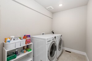 A laundry room with a front-loading washer and dryer, shelves with cleaning supplies, and a vent on the wall. - Westward Landing | Furnished Rental, 2-Car Garage (Greeley)