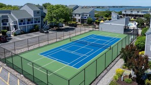 A tennis court surrounded by residential buildings, a parking area, and a view of a body of water. - East of the Sun 39866 E Sun Dr 327 (Fenwick Isle)