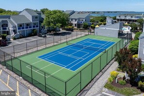 Ein Tennisplatz, umgeben von Wohngebäuden, einem Parkplatz und Blick auf ein Gewässer.